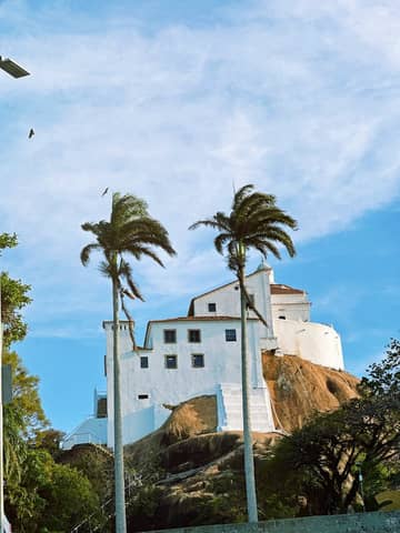 White house on hill with palm trees under blue sky, evokes tropical serenity.
