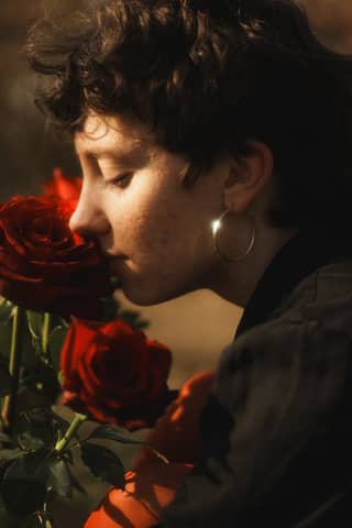 A young woman with short hair gently smells blossoming red roses outdoors.