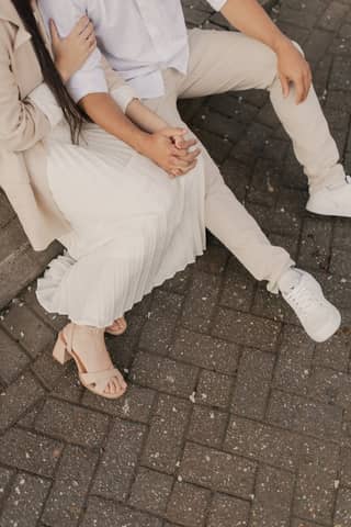 Stylish young couple sitting outdoors on paved ground wearing beige and white casual attire.