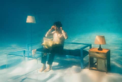 Person in an underwater living room setting with sofa and lamps.