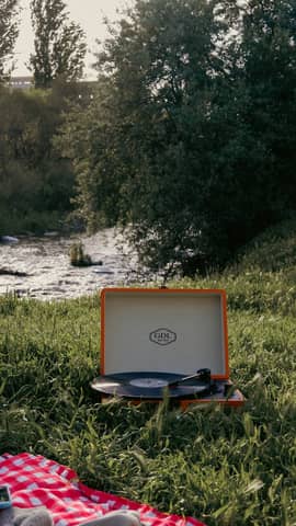 Retro record player set on a picnic blanket by a peaceful riverside, surrounded by lush greenery.