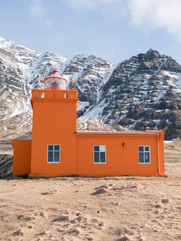 An orange lighthouse stands boldly against snow-capped mountains, creating a stunning contrast.