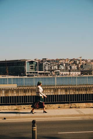 A stylish woman strolls along the riverbank with a vibrant cityscape in the background.
