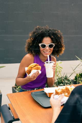 A woman sitting at a table eating a sandwich and drinking a drink