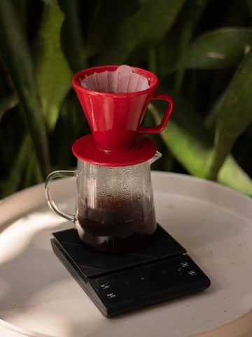 Red filter cone on glass carafe brews fresh coffee over digital scale.