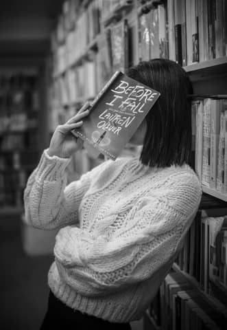 Black and white portrait of a woman hiding behind a book in a library setting.