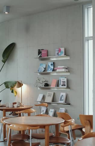 A stylish modern cafe interior featuring a wall bookshelf, wooden tables, and a potted plant.