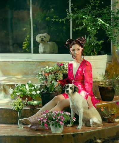 A woman in a red dress poses with dogs and potted plants in a lush garden setting.