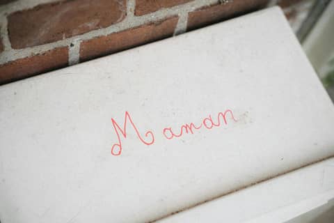 Close-up of red handwritten 'Maman' on a white concrete surface against a brick wall.