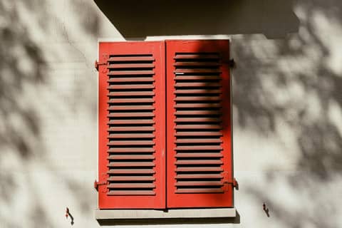 Bright red wooden shutters casting shadows on a sunlit wall in Kassel, Germany.