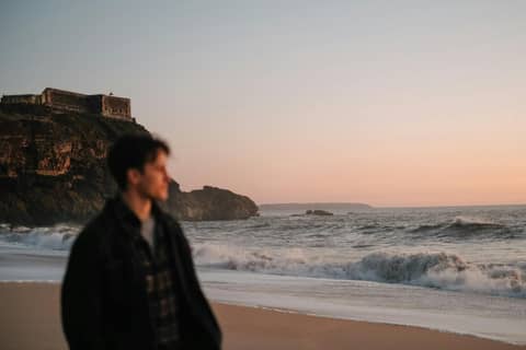 An adult male stands on a beach at sunset near coastal cliffs and ocean waves.
