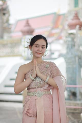 Smiling woman in ornate Thai dress posing at a temple with a traditional gesture.