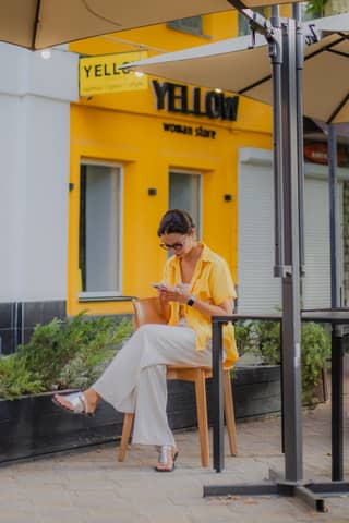 A woman wearing a yellow shirt sits outside a cafe, focusing on her smartphone.