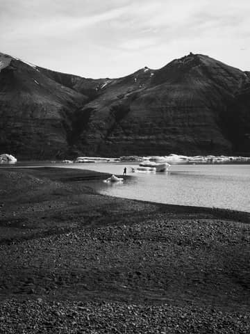 Black and white photo of a mountainous landscape with a person near an iceberg in a lake.