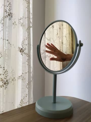 A hand reflected in a round mirror on a wooden table with light curtains in a minimalist room.
