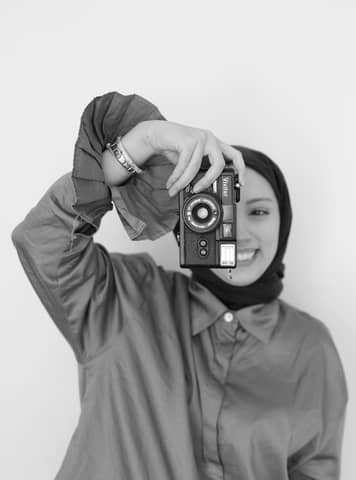 Smiling woman in hijab holds vintage camera in monochrome.