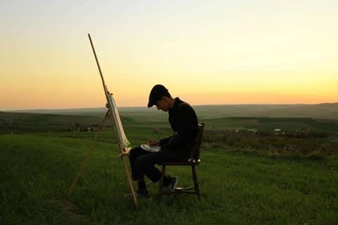 Artist paints on canvas in a peaceful field during a sunset, capturing the serene landscape.