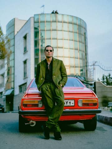 Man in green suit leaning on a classic red car in an urban setting.