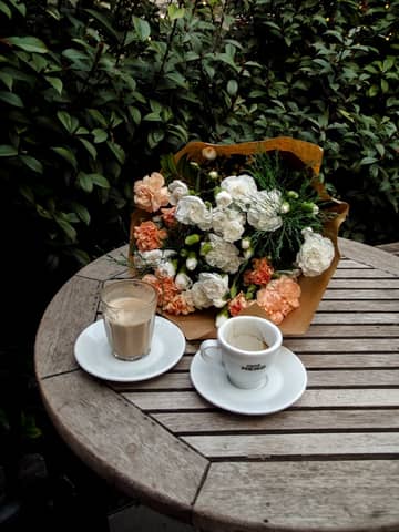 Wooden table with coffee cups and a bouquet, set in a lush garden setting.