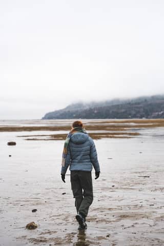A lone woman walks on a misty, serene beach with distant mountains.