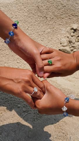 Three adults' hands adorned with colorful jewelry, signifying friendship and unity on a sandy beach.