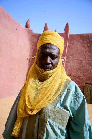 Portrait of man wearing vibrant traditional attire against adobe wall in desert landscape.