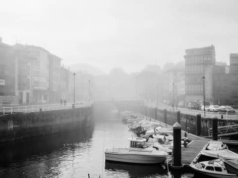 Serene black and white image of boats along a foggy riverside with bridge.